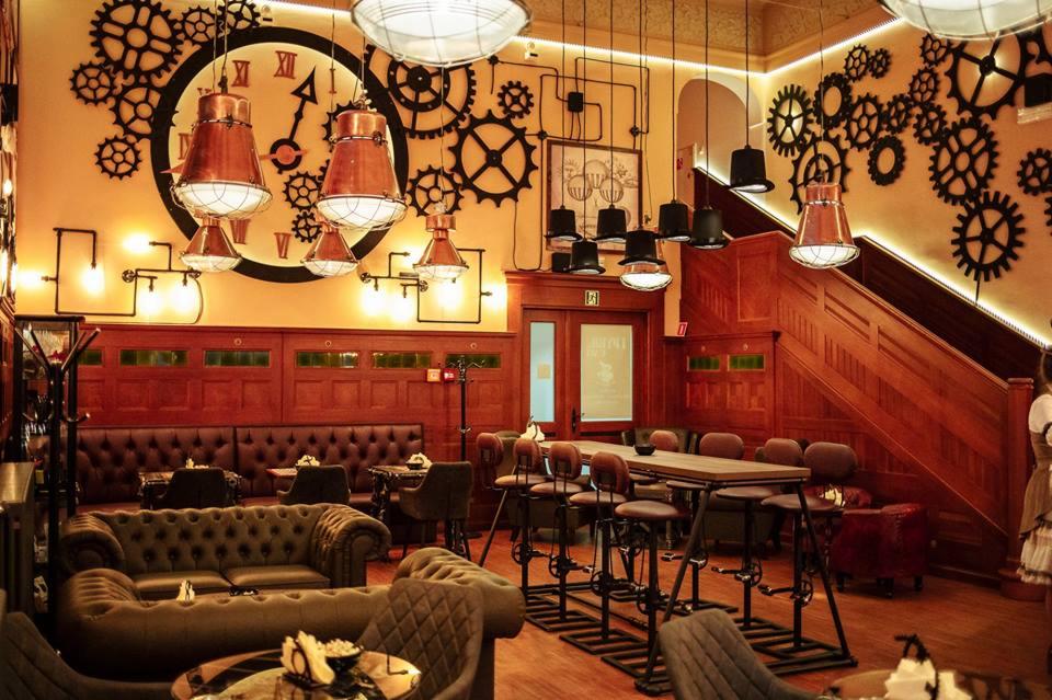 Wnętrze lokalu w stylu steampunk: drewniane panele, sofa Chesterfield, wysokie krzesła barowe z motywem przekładni, dekoracje ścienne z zębatek i zegara, miedziane lampy wiszące.