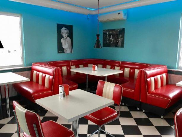 Wnętrze restauracji w stylu retro z czerwonymi lożami i krzesłami z białymi akcentami, białymi stolikami i czarno-białą podłogą w szachownicę, oświetlone niebieskim światłem.
