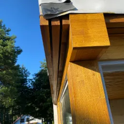 Fragment domu w konstrukcji szkieletowej z widoczną drewnianą ramą okienną, zabezpieczony od góry białą plandeką, w tle drzewa i dom.