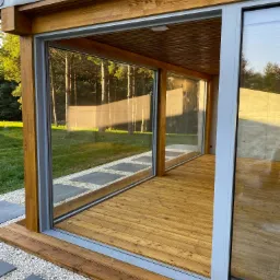 Nowoczesna weranda z drewnianą podłogą i dużymi przesuwnymi oknami, widok na ogród z drzewami w tle, ścieżka z kamiennych płyt prowadzi do wejścia, jasne naturalne światło.