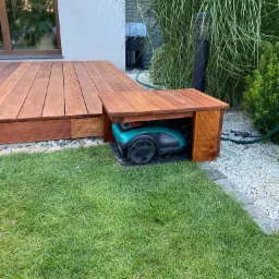 Nowoczesny taras z drewna egzotycznego z zadaszeniem, pod kt&oacute;rym schowany jest robot koszący. Otoczenie tarasu to trawnik, białe kamienie i wysokie trawy ozdobne.