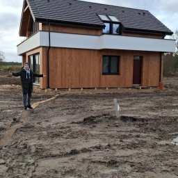 EZ CORP. SPÓŁKA Z OGRANICZONĄ ODPOWIEDZIALNOŚCIĄ - Nowoczesny dom z drewnianą elewacją i białym pasem na piętrze, osoba z rozłożonymi rękami na tle błotnistego terenu.