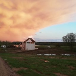 EZ CORP. SPÓŁKA Z OGRANICZONĄ ODPOWIEDZIALNOŚCIĄ - Wznoszenie domu w Częstochowie na tle dramatycznego nieba. Widoczny szkielet budynku z dachem, otoczony zielenią i zbiornikiem wodnym w oddali.