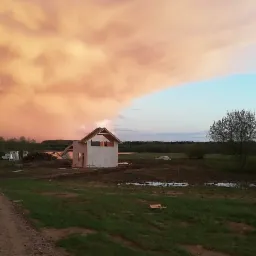 Wznoszenie domu w Częstochowie na tle dramatycznego nieba. Widoczny szkielet budynku z dachem, otoczony zielenią i zbiornikiem wodnym w oddali.