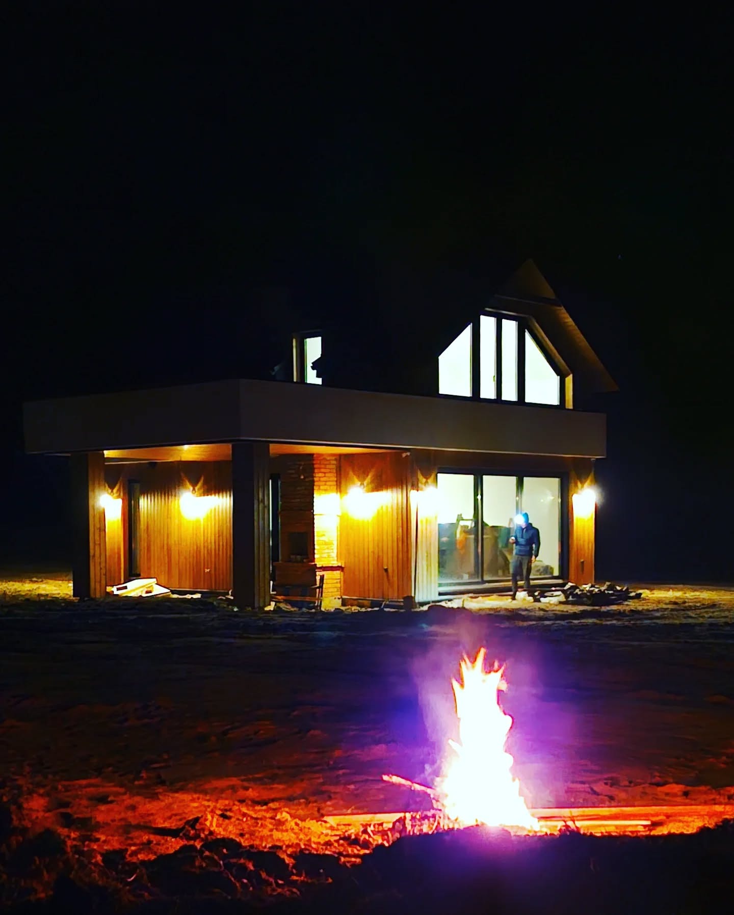Nocna sceneria z płonącym ogniskiem przed nowoczesnym domem z drewnianą elewacją i oświetlonymi oknami, osoba stoi przed wejściem.