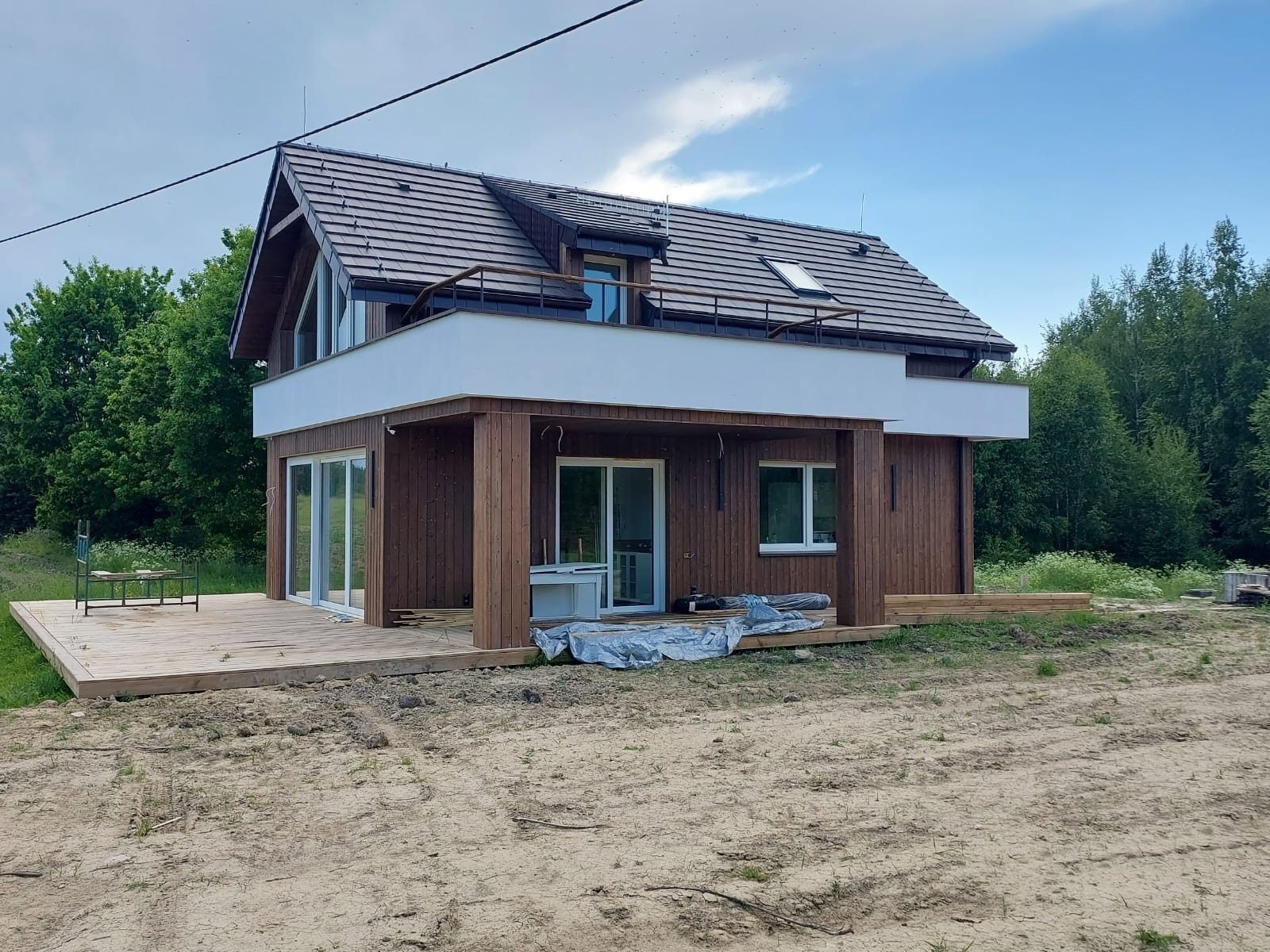 Nowoczesny dom z drewnianą elewacją i tarasem w trakcie budowy, z widocznymi elementami wykończeniowymi i otaczającą zielenią.