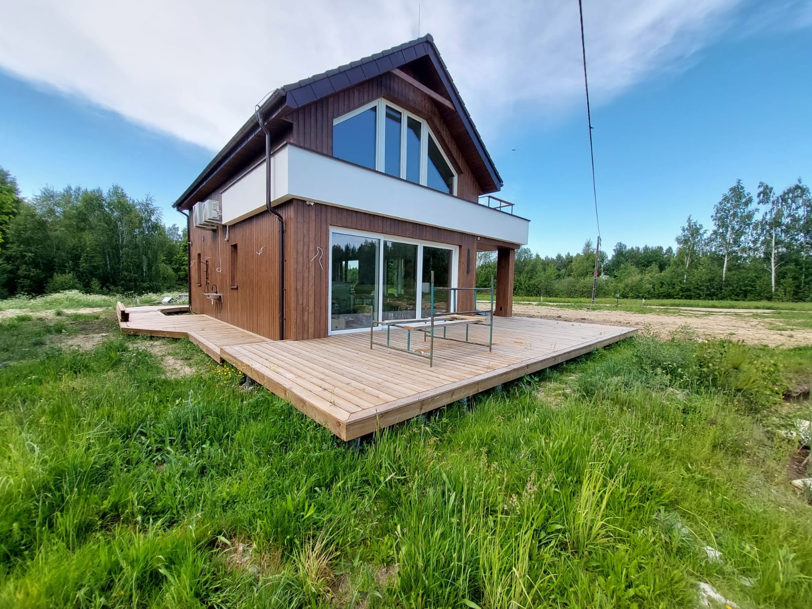 Nowoczesny dom z drewnianą elewacją i tarasem z jasnego drewna, otoczony zielenią. Widoczne duże przesuwne okna i balkon z białą balustradą.