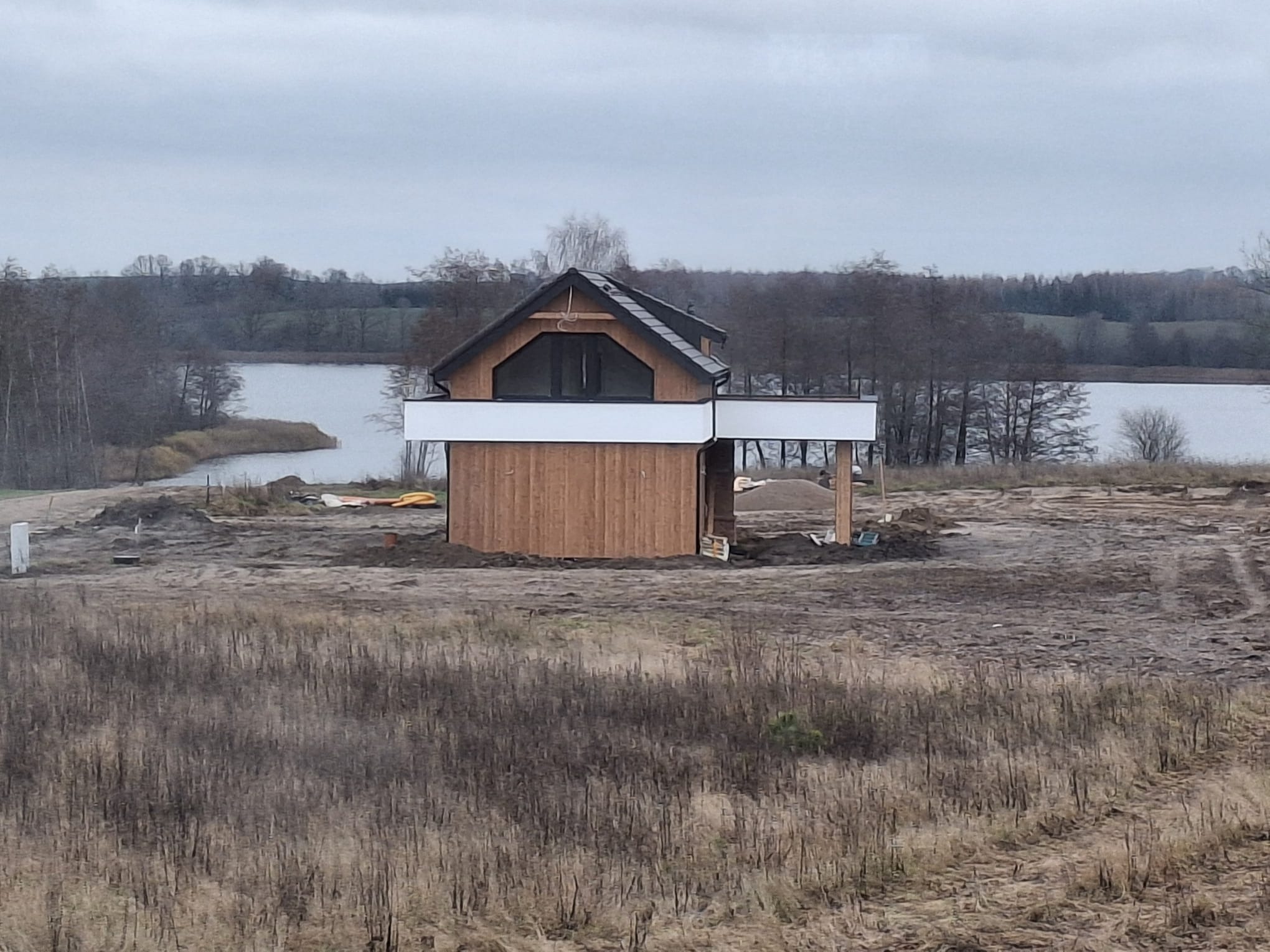 Niedokończony, drewniany dom z białym tarasem na wzgórzu, z widokiem na jezioro w pochmurny dzień. Widoczne materiały budowlane i teren w trakcie przygotowywania.