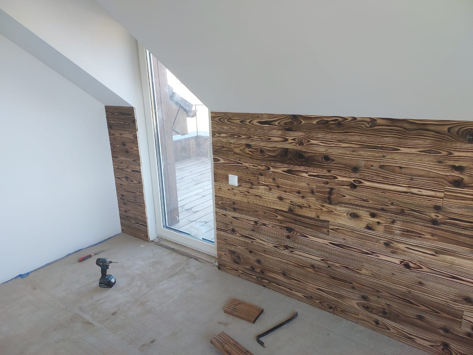 Montaż opalanej boazerii na ścianie poddasza, widoczny fragment okna, narzędzia i deski. Detal wykończenia wnętrza z unikalnym wzorem drewna.
