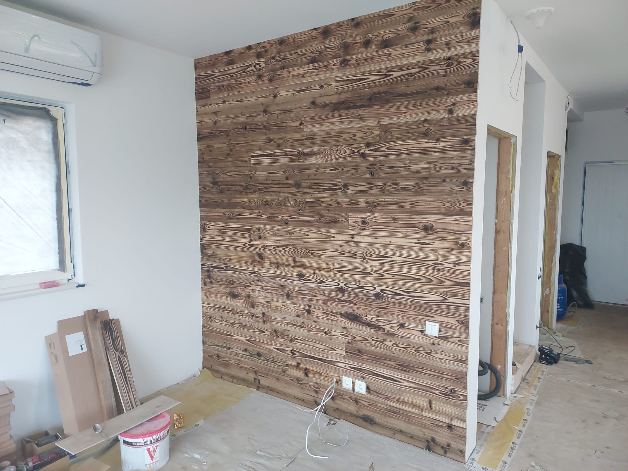 Drewniana ściana z desek o wyraźnym rysunku słojów w jasnych i ciemnych odcieniach brązu, zamontowana w nowym wnętrzu. Widoczne gniazdka elektryczne i elementy wykończeniowe.