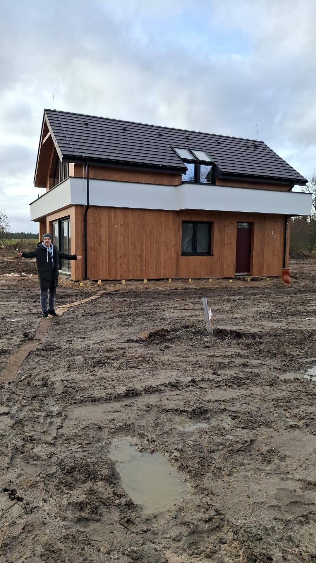 Nowoczesny dom z drewnianą elewacją i białym pasem na piętrze, osoba z rozłożonymi rękami na tle błotnistego terenu.