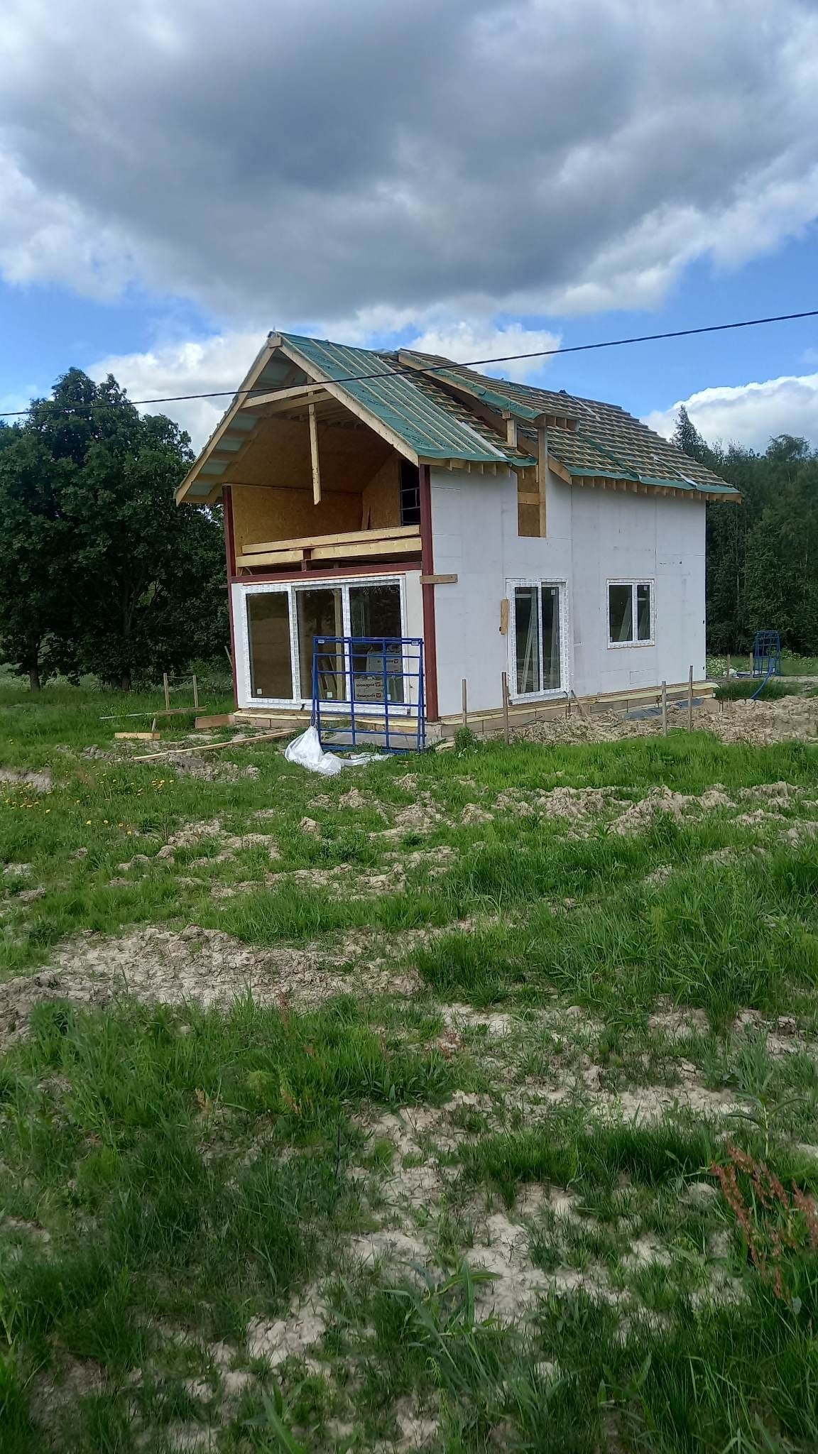 Budowa domu szkieletowego w toku: widoczne ściany z płyt OSB, częściowo pokryty dach z zieloną membraną, okna PCV i rusztowanie na trawiastym terenie.