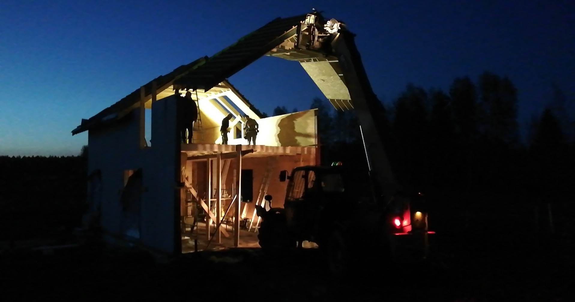 Budowa domu szkieletowego w nocy: ekipa montuje elementy konstrukcyjne przy użyciu dźwigu teleskopowego. Widoczna konstrukcja, drewniane elementy i sylwetki robotników.