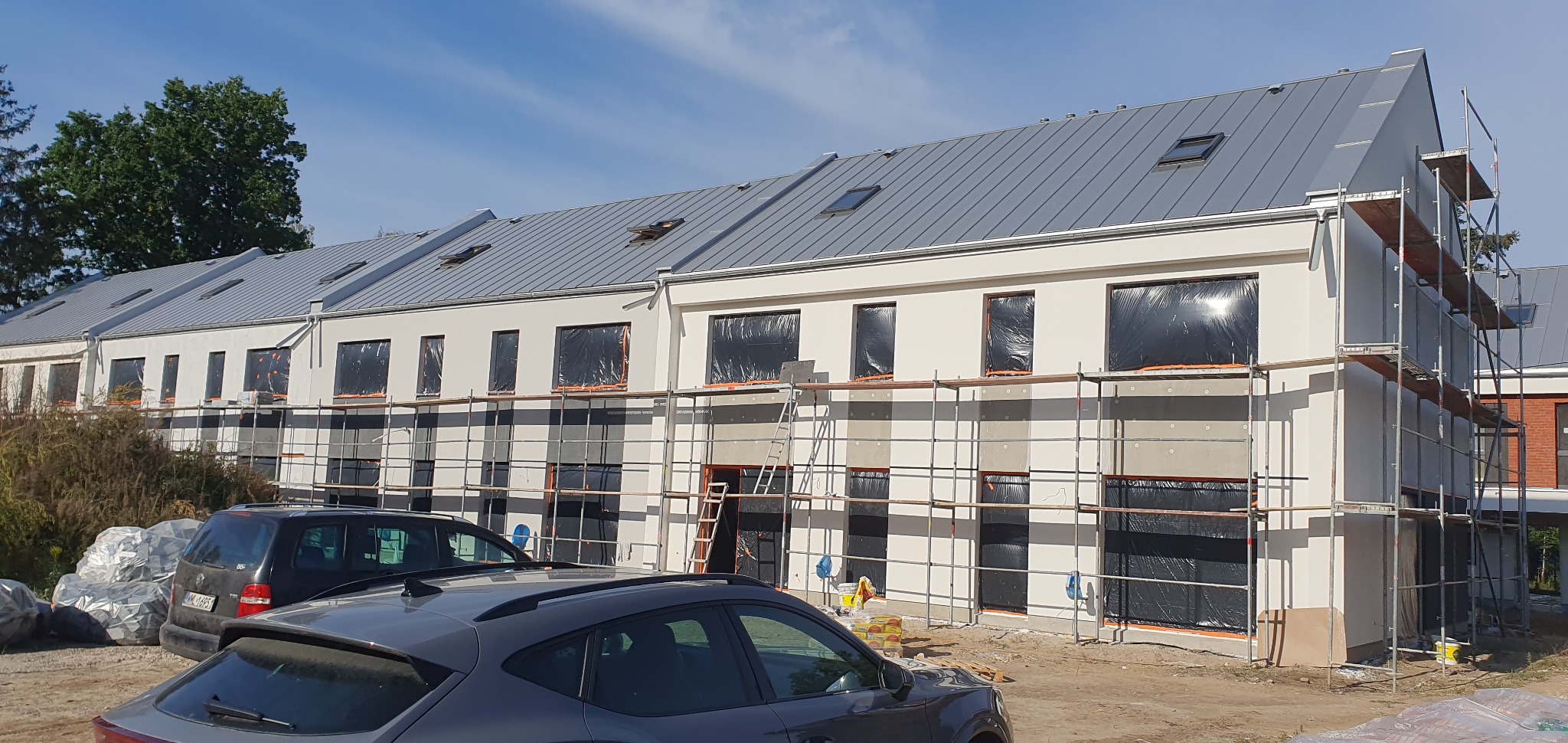 Szeregowy budynek mieszkalny w trakcie ocieplania, okna zabezpieczone folią, rusztowania ustawione wzdłuż elewacji, na pierwszym planie zaparkowane samochody.