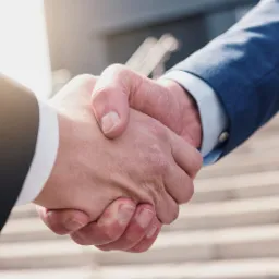 Uścisk dłoni dwóch mężczyzn w garniturach na tle schodów budynku.