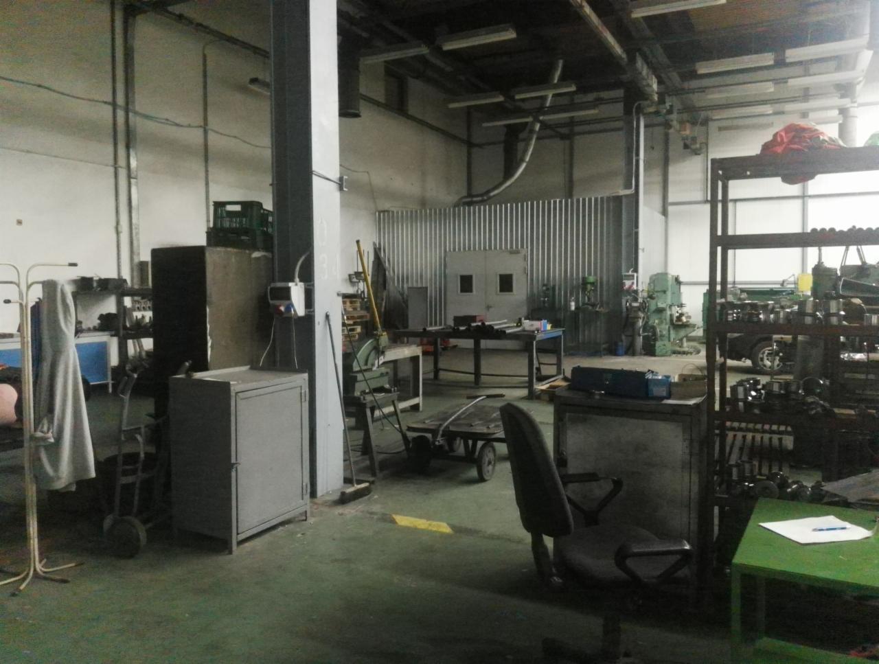 Wnętrze warsztatu metalurgicznego z różnorodnym sprzętem, w tym maszynami do obróbki metalu, regałami z elementami metalowymi i wózkiem transportowym. Widoczna metalowa ściana z drzwiami...