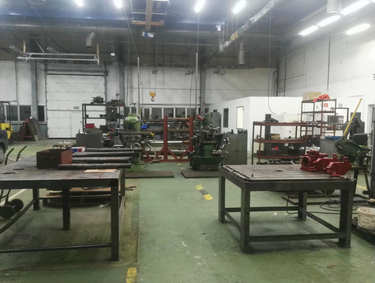 Warsztat obróbki metali: stoły robocze z metalowymi elementami, w tle tokarki i inne maszyny, suwnica pod sufitem.