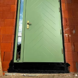 Świeżo zamontowane, pistacjowe drzwi wejściowe z pionowym przeszkleniem, widoczne czarne uszczelnienie wokół ramy i mur z cegły ceramicznej.