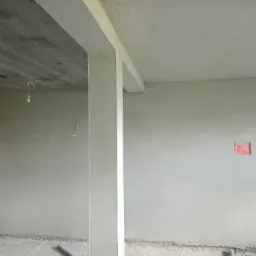 Wnętrze w trakcie prac wykończeniowych: ściany pokryte świeżym tynkiem, widoczny surowy betonowy strop i niedokończone instalacje elektryczne.