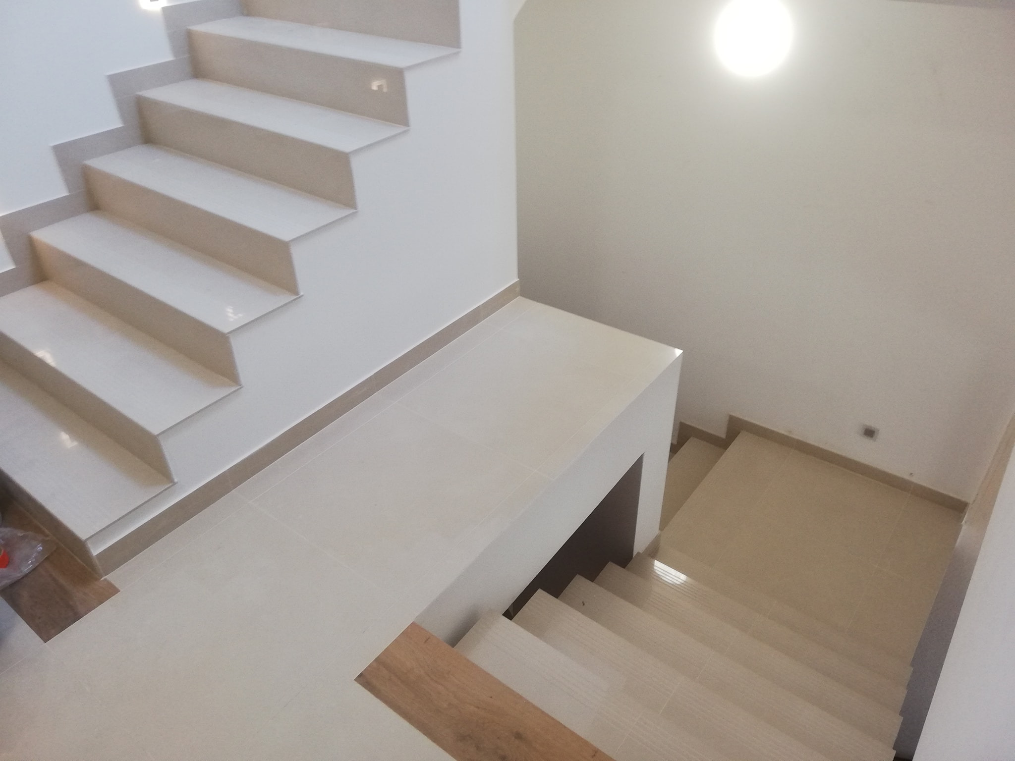 Wykończone schody z jasnymi, beżowymi płytkami ceramicznymi na stopniach i podłodze, widok z góry na dół, z oświetleniem punktowym w suficie.