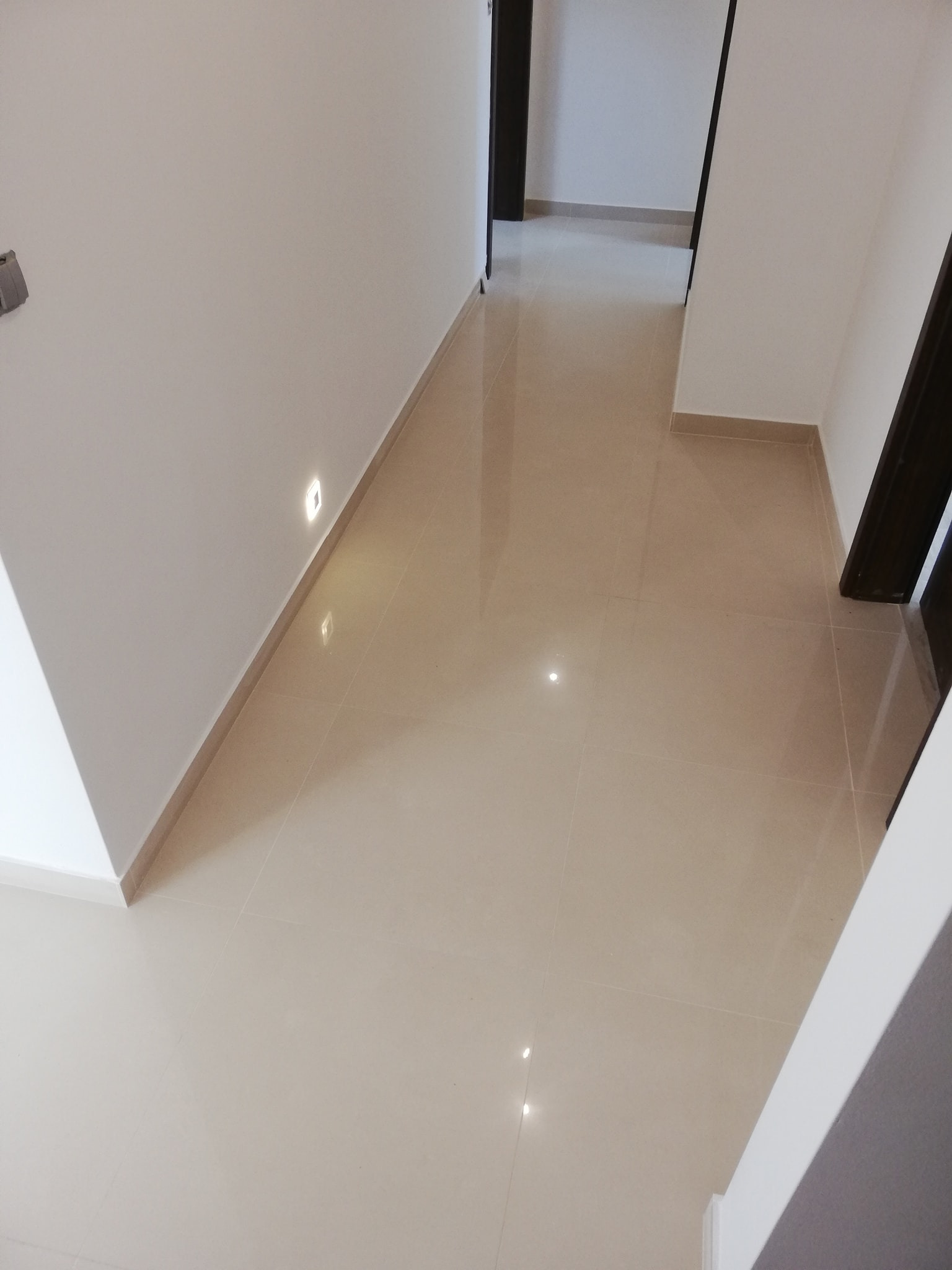 Beżowe, błyszczące płytki podłogowe w korytarzu z wbudowanym oświetleniem LED przy ścianie i widocznymi, ciemnymi futrynami drzwi.