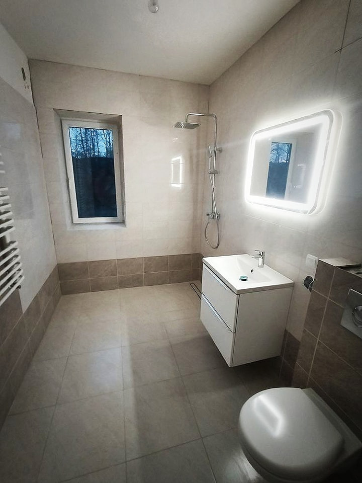 Wykończona łazienka z beżowymi płytkami, podświetlanym lustrem i kabiną prysznicową typu walk-in, widoczne okno z widokiem na drzewa.