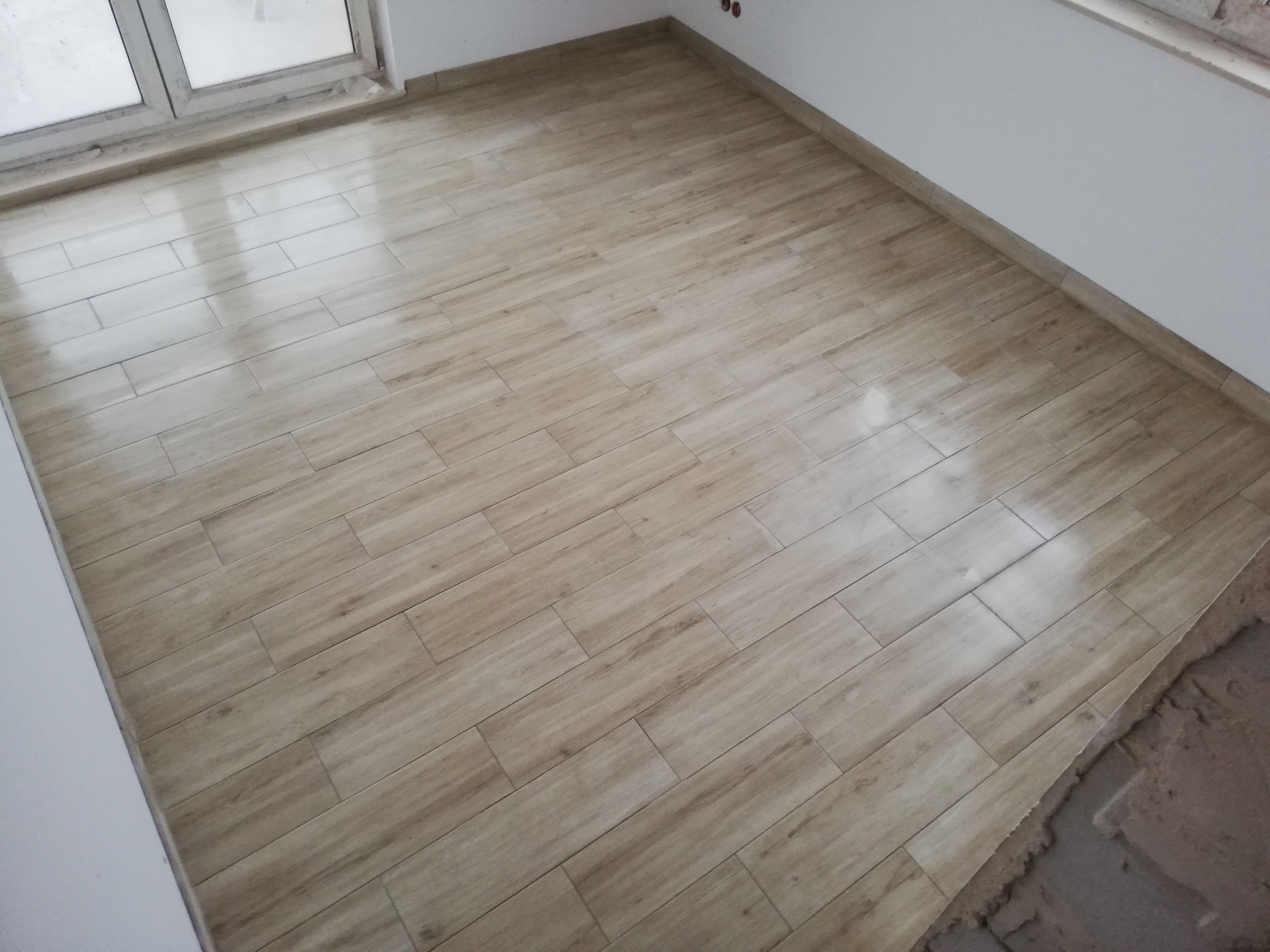 Ułożona podłoga z płytek imitujących drewno w jasnym odcieniu, widoczne fugi, fragment okna i surowy beton w rogu pomieszczenia.