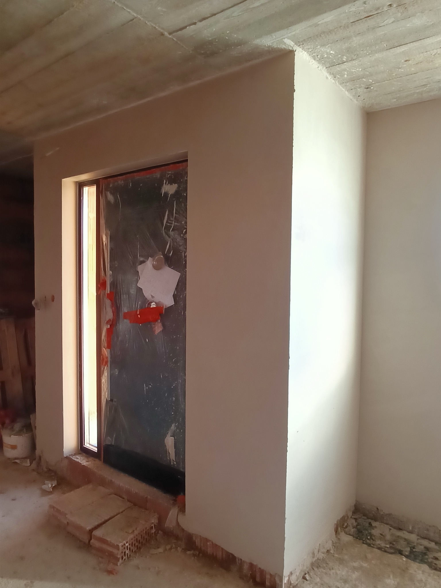 Świeżo położone, gładkie tynki maszynowe na ścianie w nowym budynku, widoczna zabezpieczona folią futryna drzwiowa i fragment surowego sufitu.