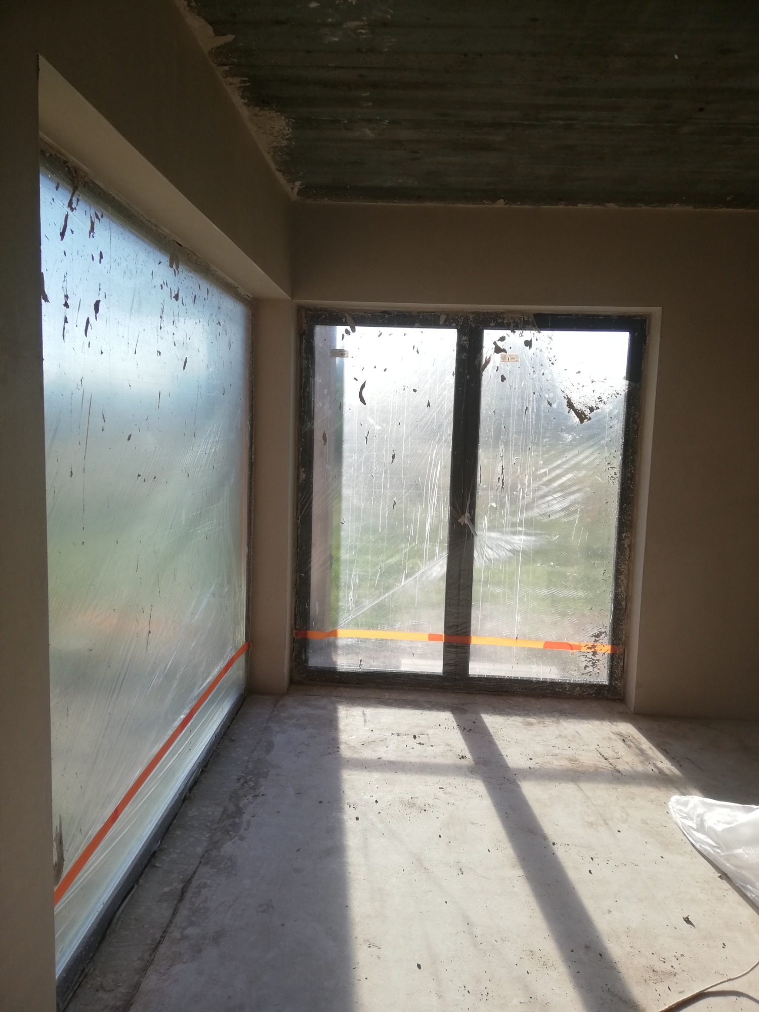 Pokój w stanie deweloperskim po wykonaniu tynków maszynowych, okna zabezpieczone folią i taśmą malarską, widoczny surowy betonowy strop.
