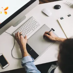 Widok z góry na biurko z komputerem, tabletem graficznym, telefonem i kubkiem kawy; ręce mężczyzny pracują na tablecie graficznym i klawiaturze, na ekranie komputera widoczna grafika z motywem...