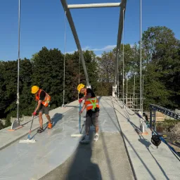 Dw&oacute;ch robotnik&oacute;w w kaskach i kamizelkach rozprowadza jasną substancję na betonowej powierzchni mostu, używając mop&oacute;w. W tle zieleń drzew i konstrukcja mostu.