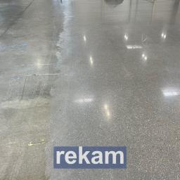 REKAM SPÓŁKA Z OGRANICZONĄ ODPOWIEDZIALNOŚCIĄ - Szlifowana, betonowa posadzka przemysłowa, z widocznym podziałem na obszar przed i po szlifowaniu, odbijająca światło lamp sufitowych.