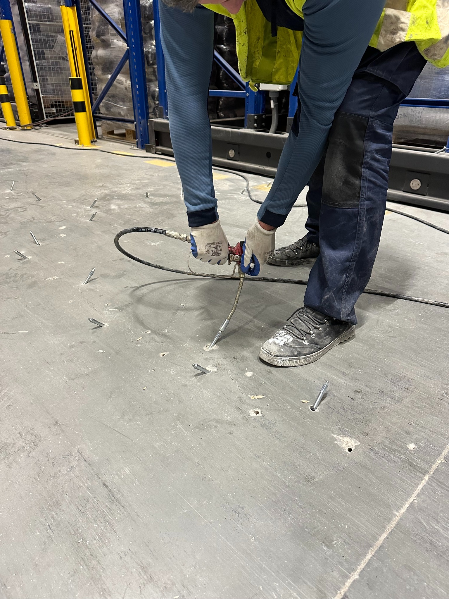 Aplikacja preparatu gruntującego na betonową posadzkę w hali magazynowej za pomocą pistoletu iniekcyjnego. Widoczne kotwy mocujące.