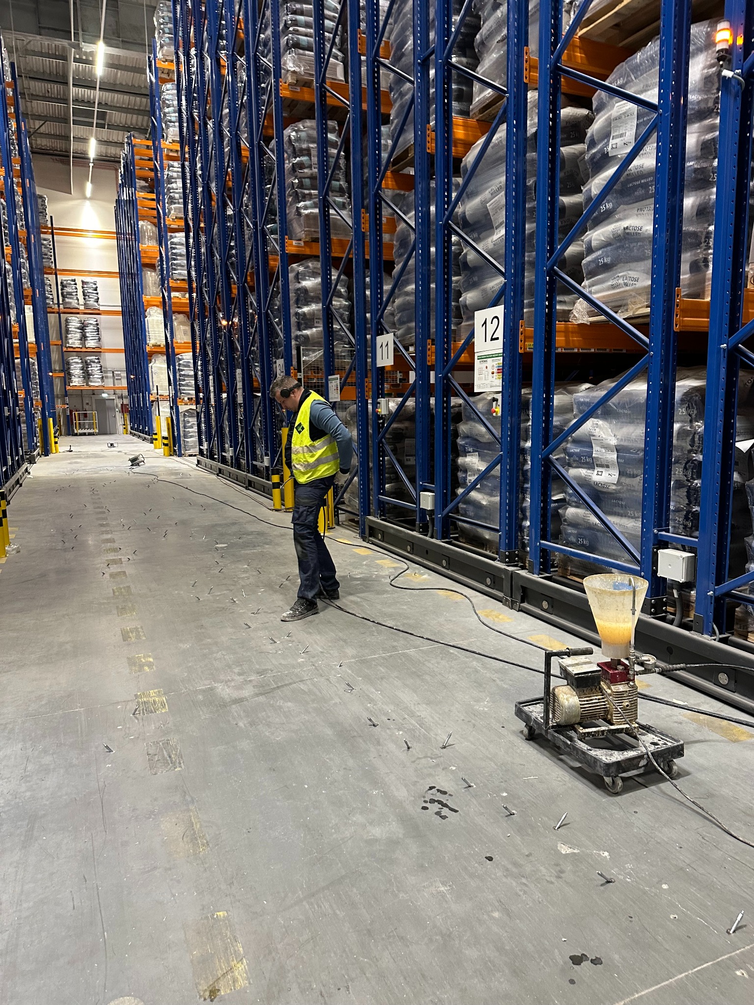 Aplikacja przemysłowej wylewki w magazynie. Pracownik w kamizelce obok maszyny do wylewania posadzki. Regały z towarem w tle. Widoczne ślady po naprawach.