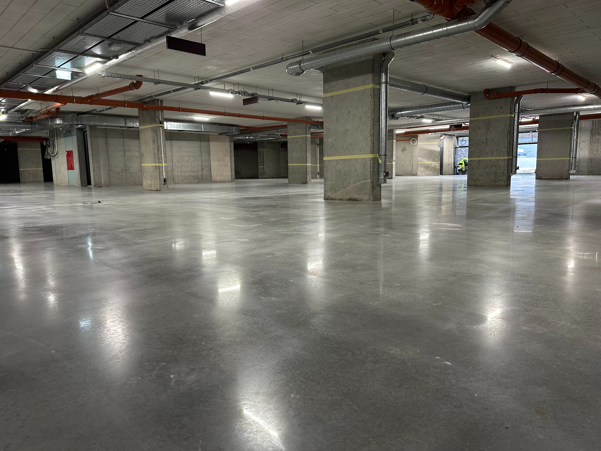 Szara, błyszcząca posadzka w hali przemysłowej z betonowymi kolumnami owiniętymi taśmą. Widoczne instalacje pod sufitem i w oddali pracownik w odblaskowej kamizelce.