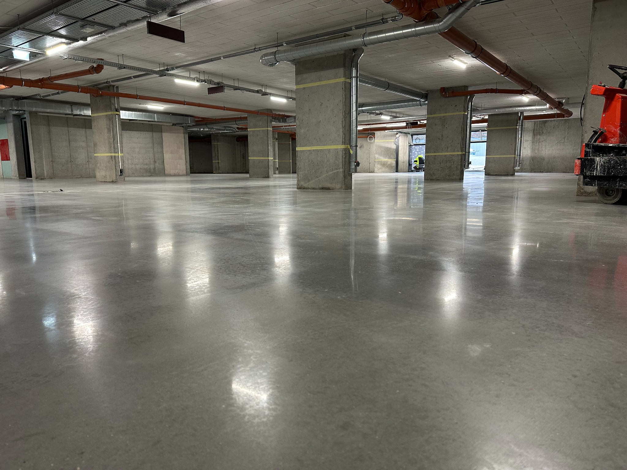 Błyszcząca, szara posadzka żywiczna w hali garażowej, z widocznymi słupami konstrukcyjnymi i instalacjami pod sufitem. Na prawo szlifierka do posadzek.