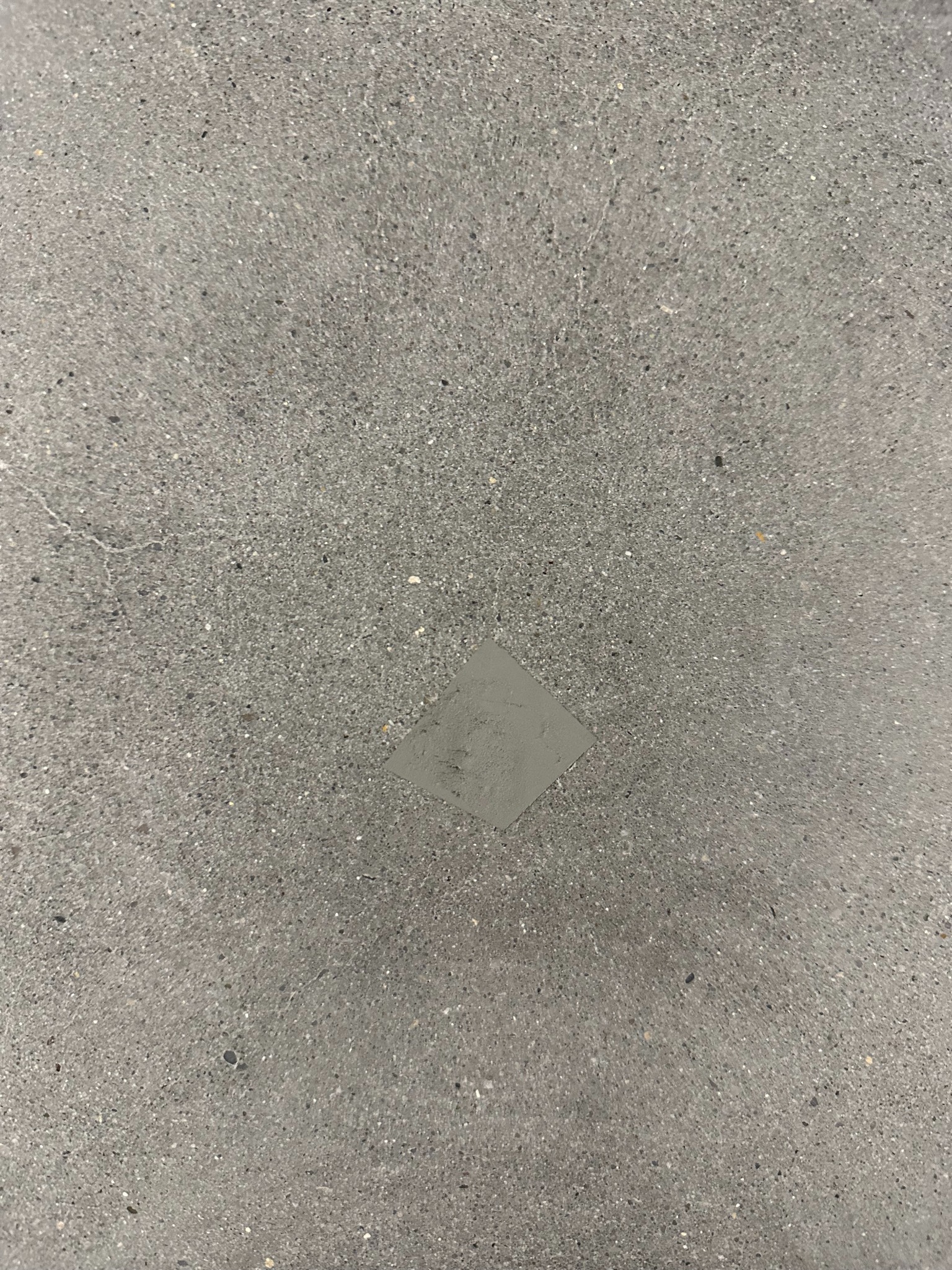 Szara posadzka z żywicy epoksydowej, z widocznym miejscem po naprawie, wypełnionym jaśniejszą żywicą. Powierzchnia lekko chropowata, matowa.