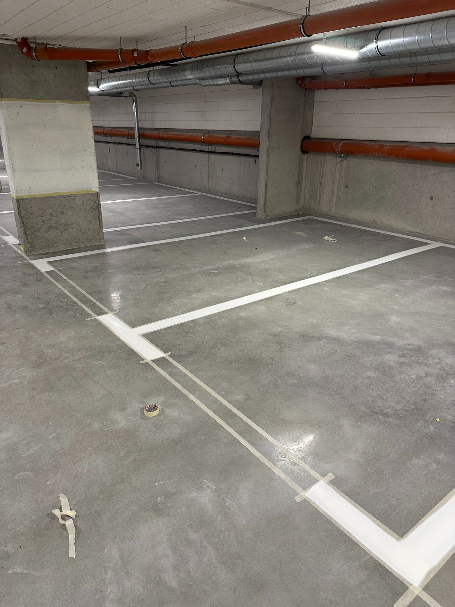 Oznakowane białą farbą stanowiska parkingowe na szarej posadzce w garażu podziemnym. Widoczne rury pod sufitem i taśma malarska.