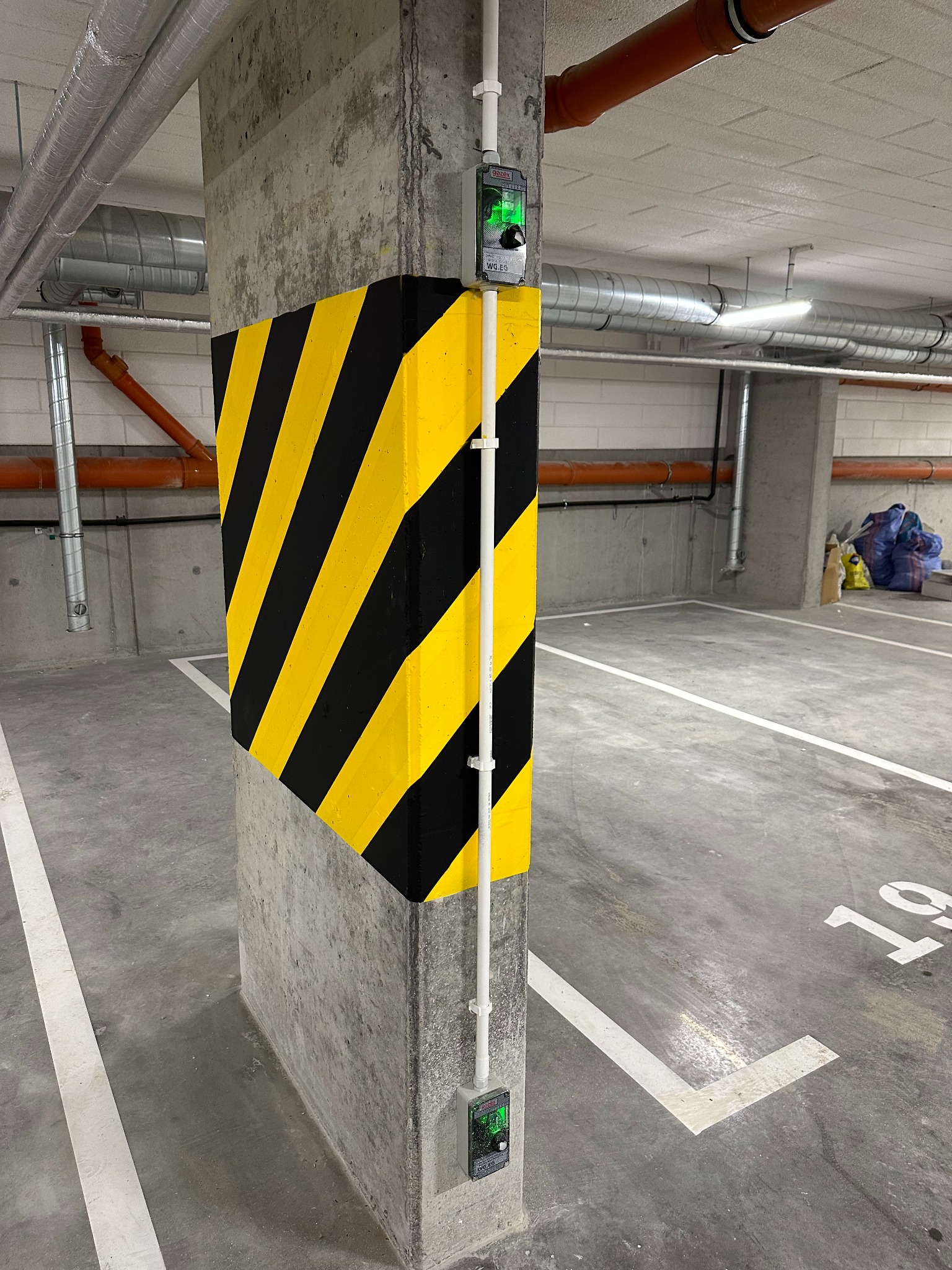Oznakowany słup w garażu podziemnym z posadzką żywiczną i instalacją elektryczną. Żółto-czarne pasy ostrzegawcze na narożniku.
