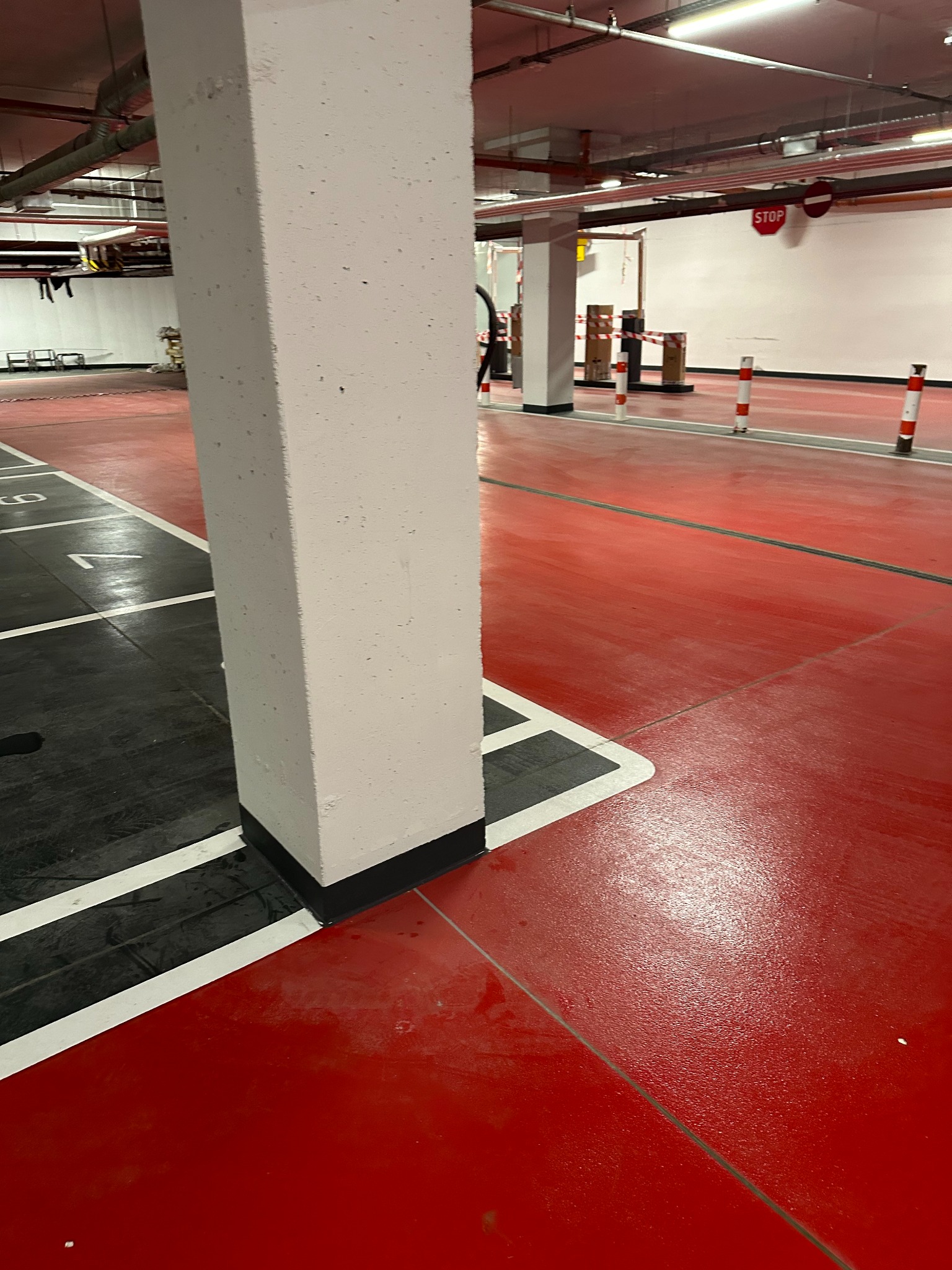 Czerwona posadzka żywiczna w garażu podziemnym z wyznaczonymi miejscami parkingowymi, widok na biały filar i oznaczenia STOP w tle.