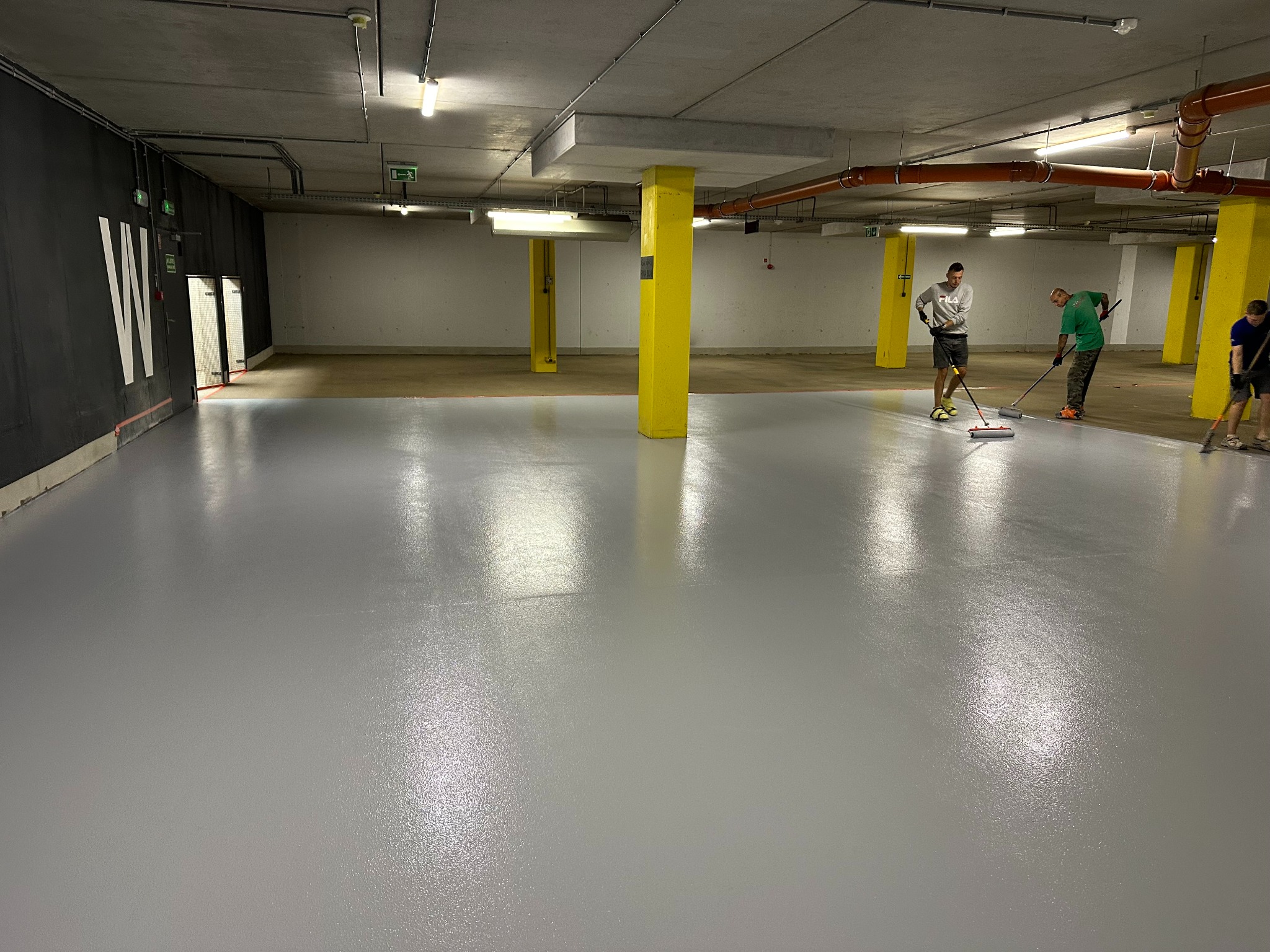 Praca wre: Trzech mężczyzn aplikuje szarą posadzkę żywiczną w przestronnym garażu z żółtymi filarami i rurami pod sufitem, używając wałków na długich kijach.