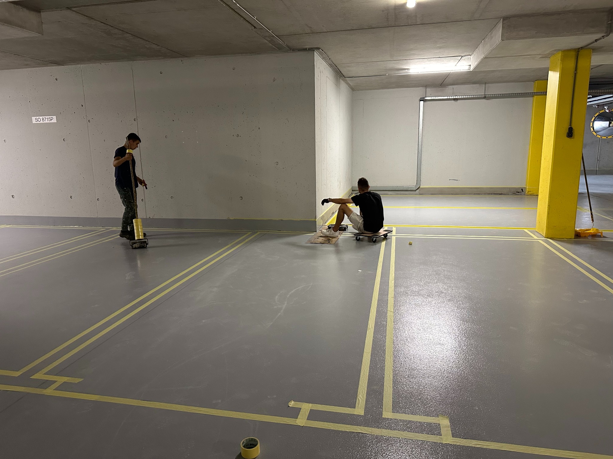 Prace wykończeniowe w garażu: dwóch mężczyzn maluje szarą posadzkę, wyznaczając żółtą taśmą granice miejsc parkingowych. Żółty słup w tle.