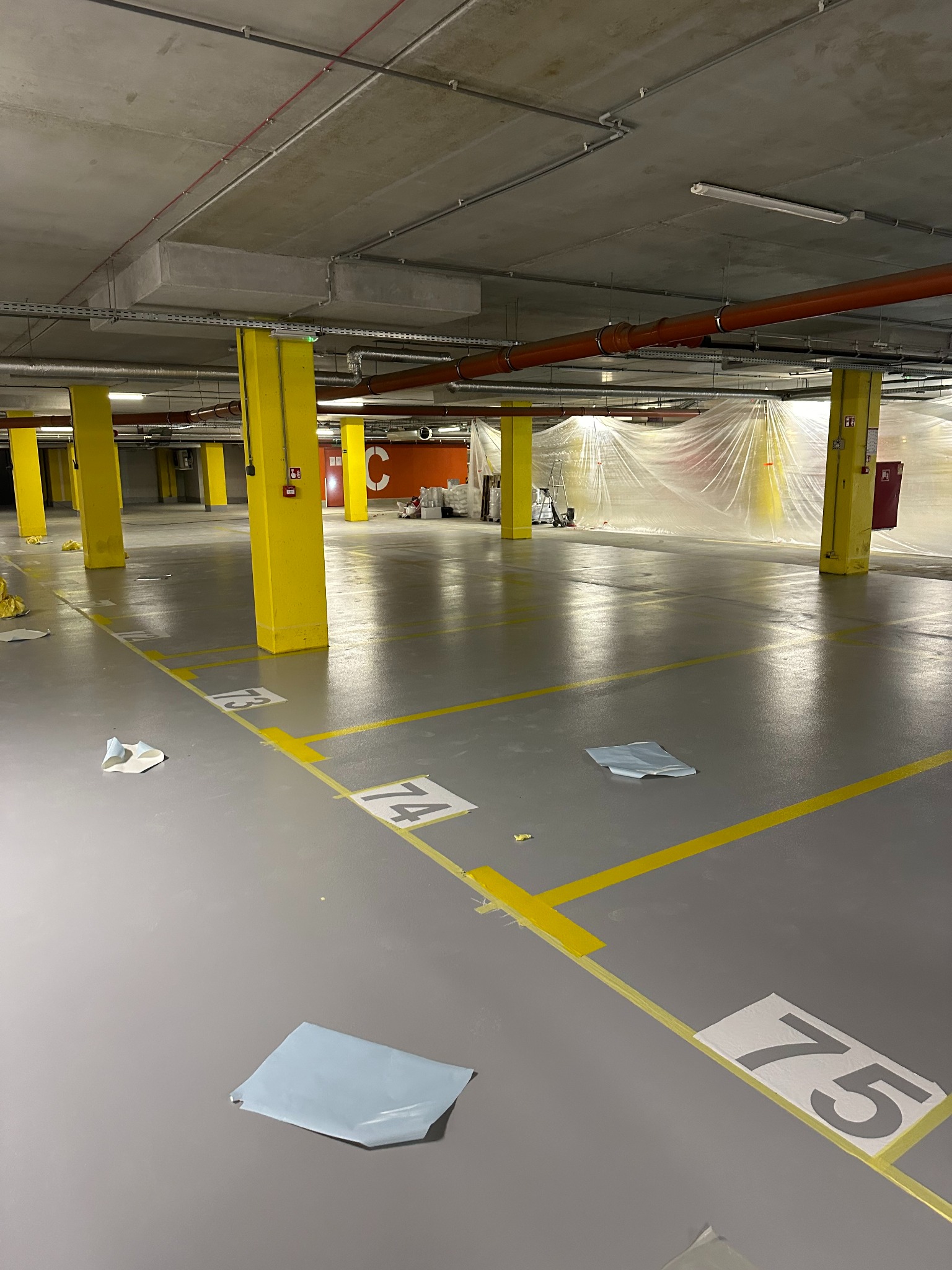 Szara posadzka w parkingu podziemnym z żółtymi słupami i numeracją miejsc. Część parkingu zabezpieczona folią malarską, widoczne niedoskonałości malowania linii.