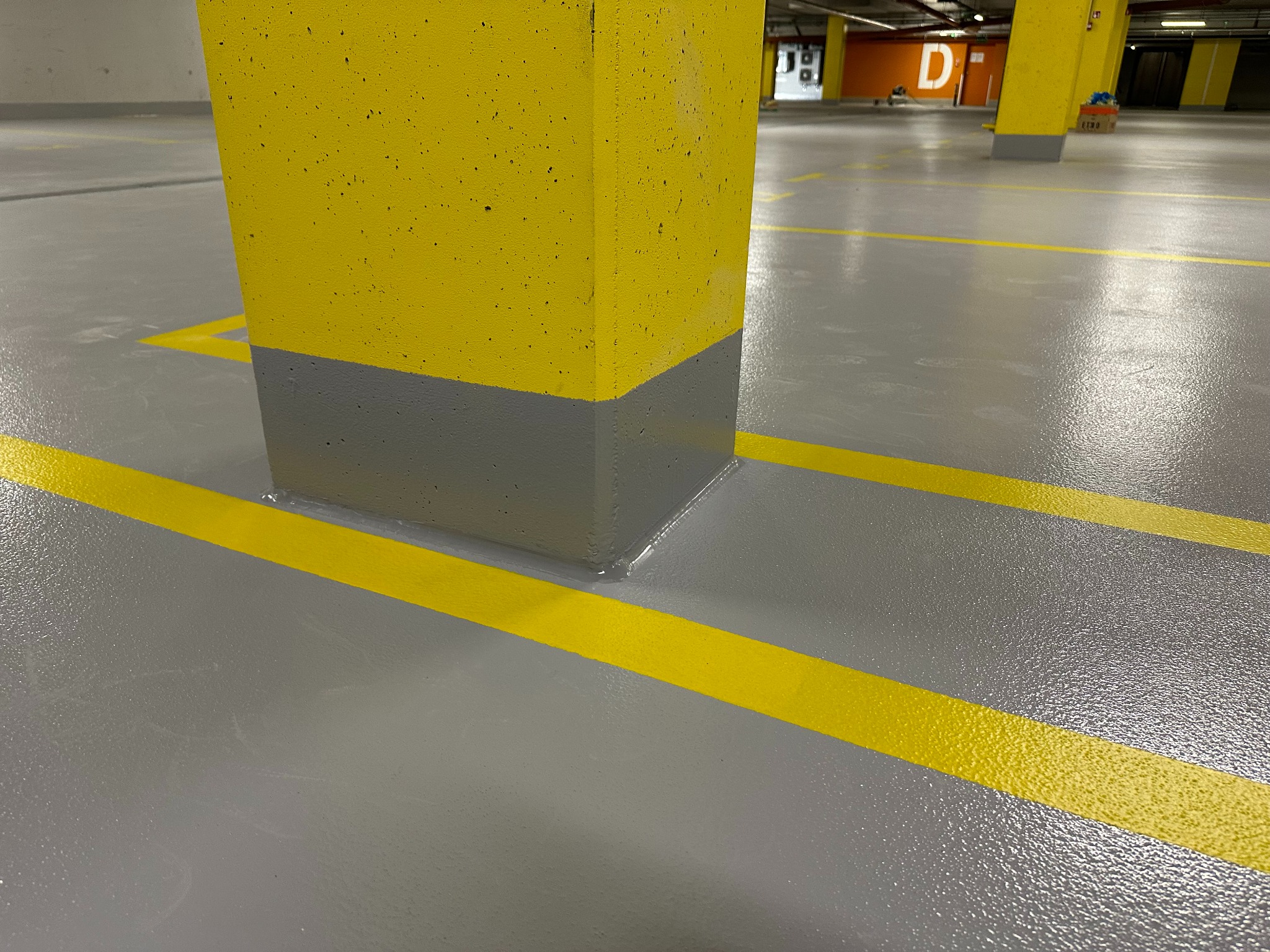 Szara posadzka żywiczna w parkingu podziemnym z żółtymi liniami i słupem w kolorze żółto-szarym. Widoczne oznaczenia stanowisk.