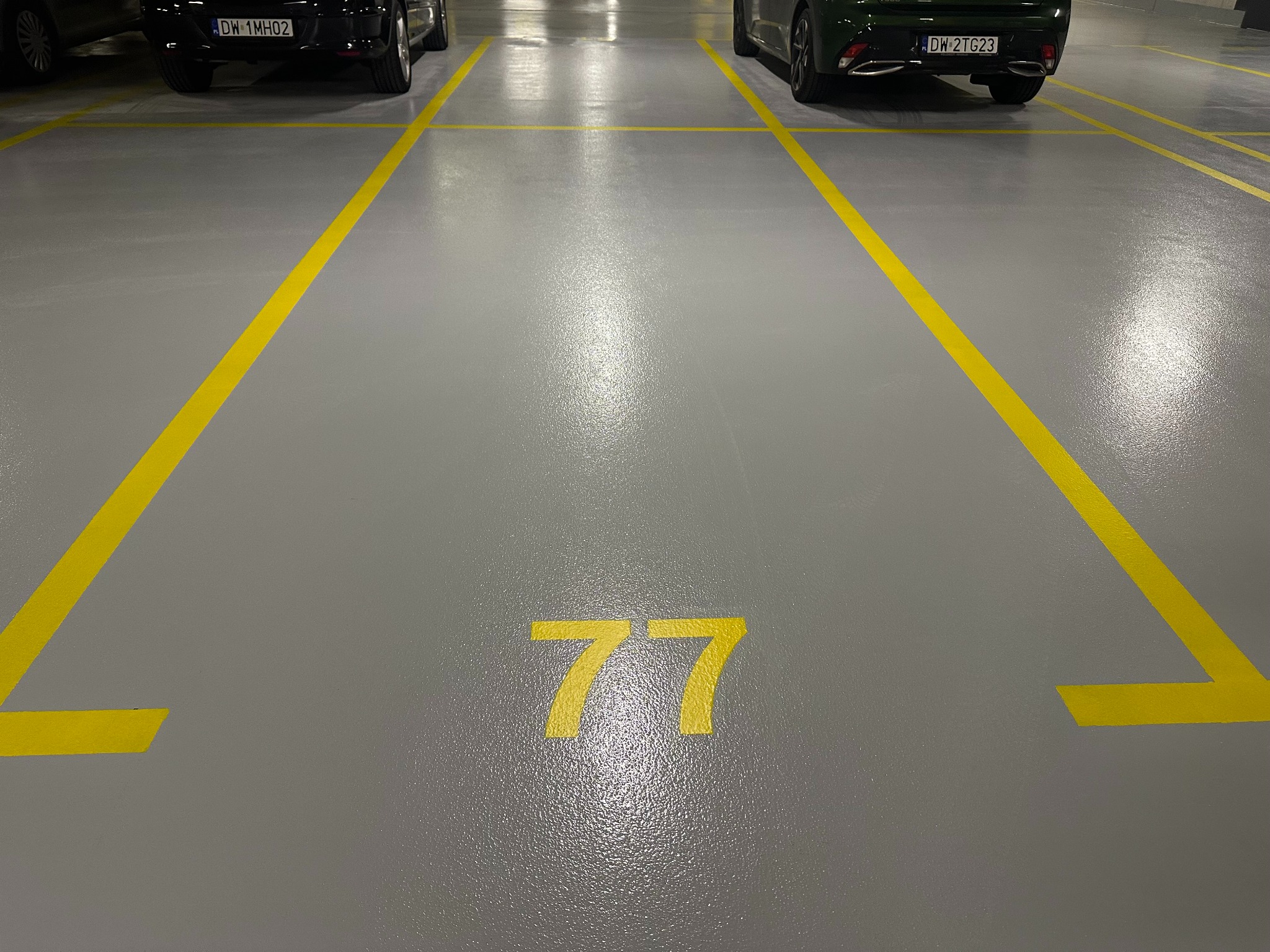 Szara posadzka żywiczna w garażu podziemnym, z żółtymi liniami wyznaczającymi miejsca parkingowe i numerem '77'. Dwa auta w tle, oświetlenie sufitowe.