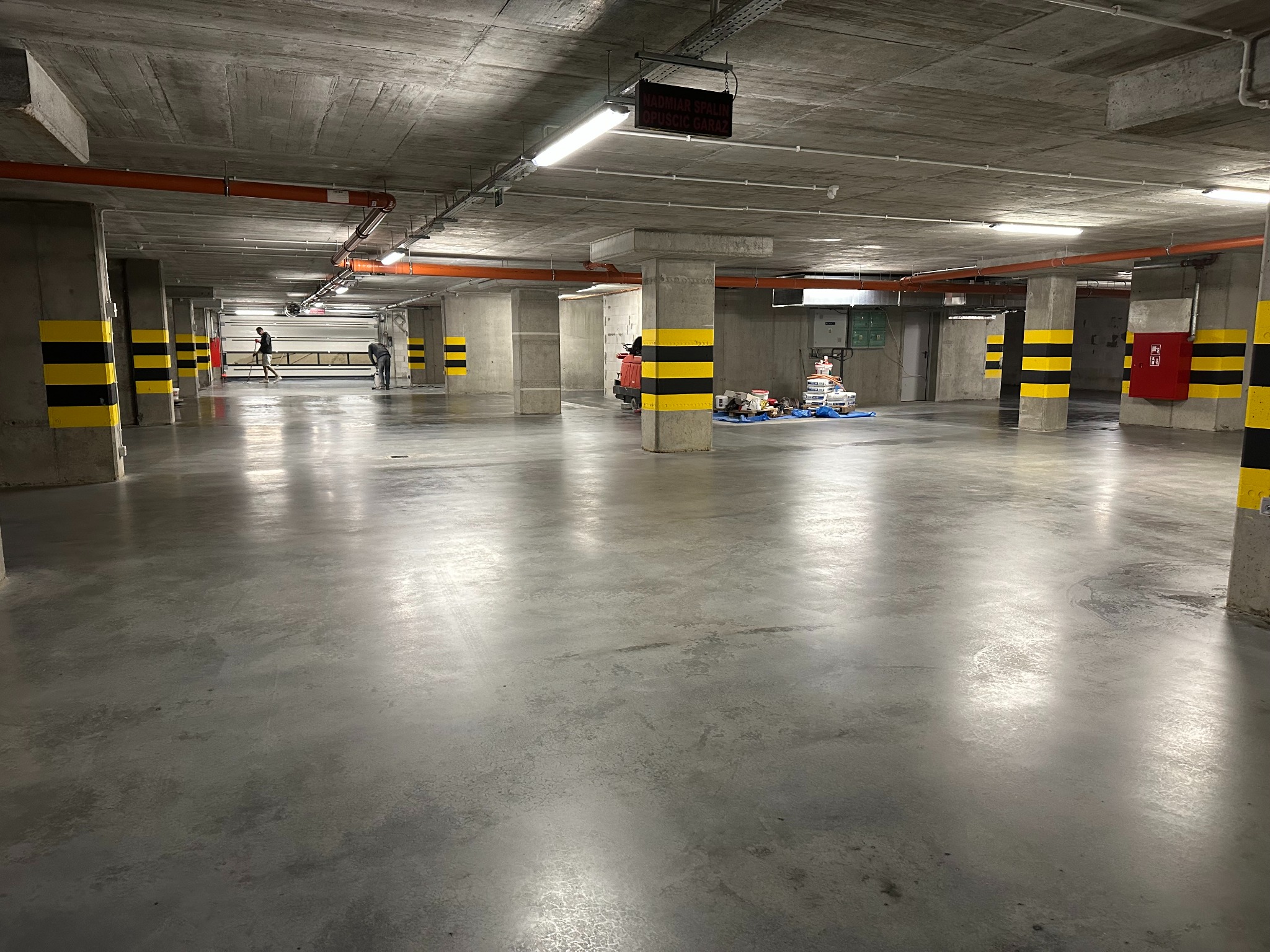 Błyszcząca, szara posadzka w garażu podziemnym, wykończona żywicą. Widoczne żółto-czarne pasy na kolumnach, sprzęt budowlany w tle i dwóch pracowników w oddali.