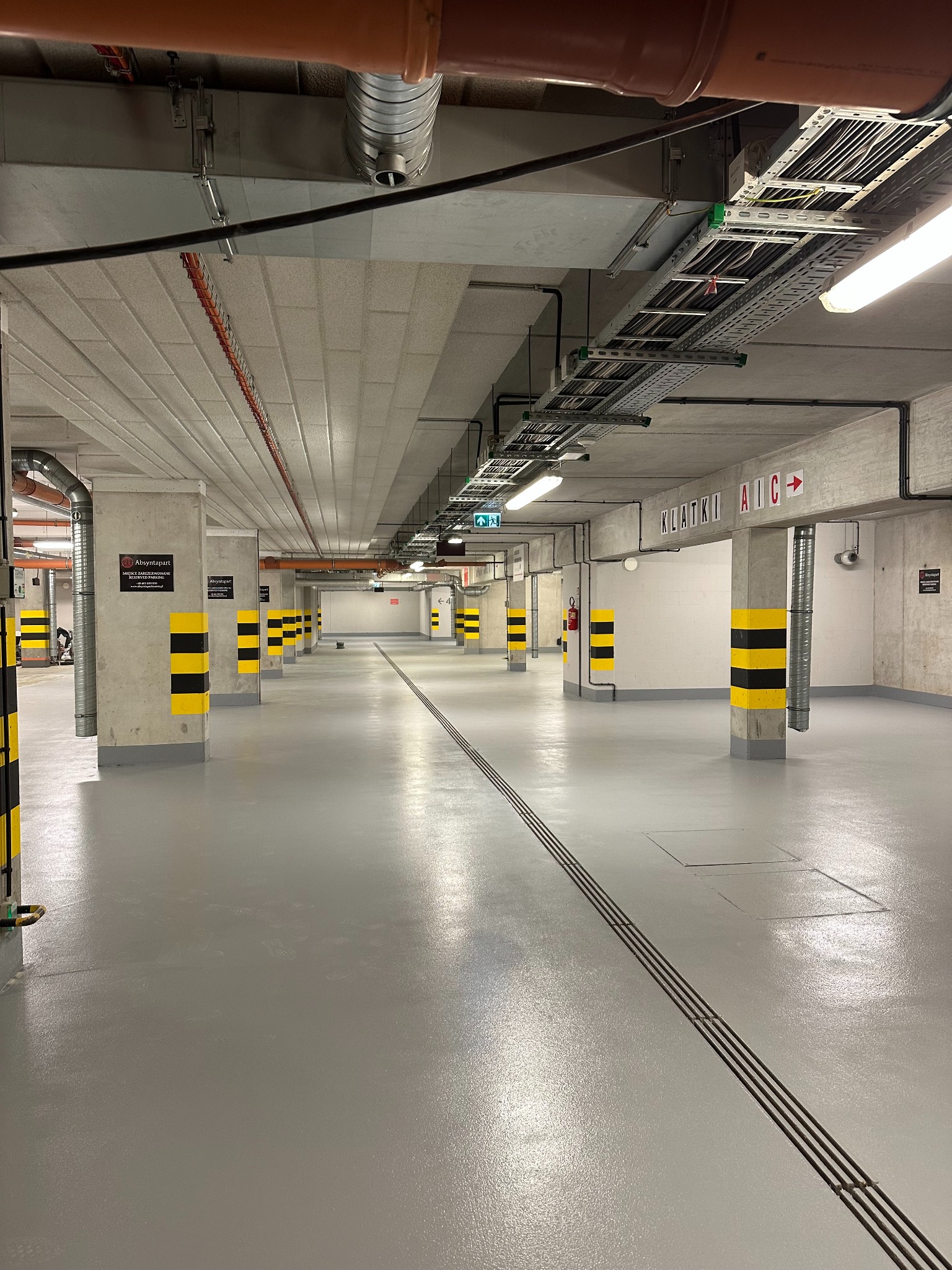 Szara, gładka posadzka żywiczna w garażu podziemnym z żółto-czarnymi pasami na kolumnach. Widoczne instalacje pod sufitem i oznaczenia 'Klatki A|B|C'.