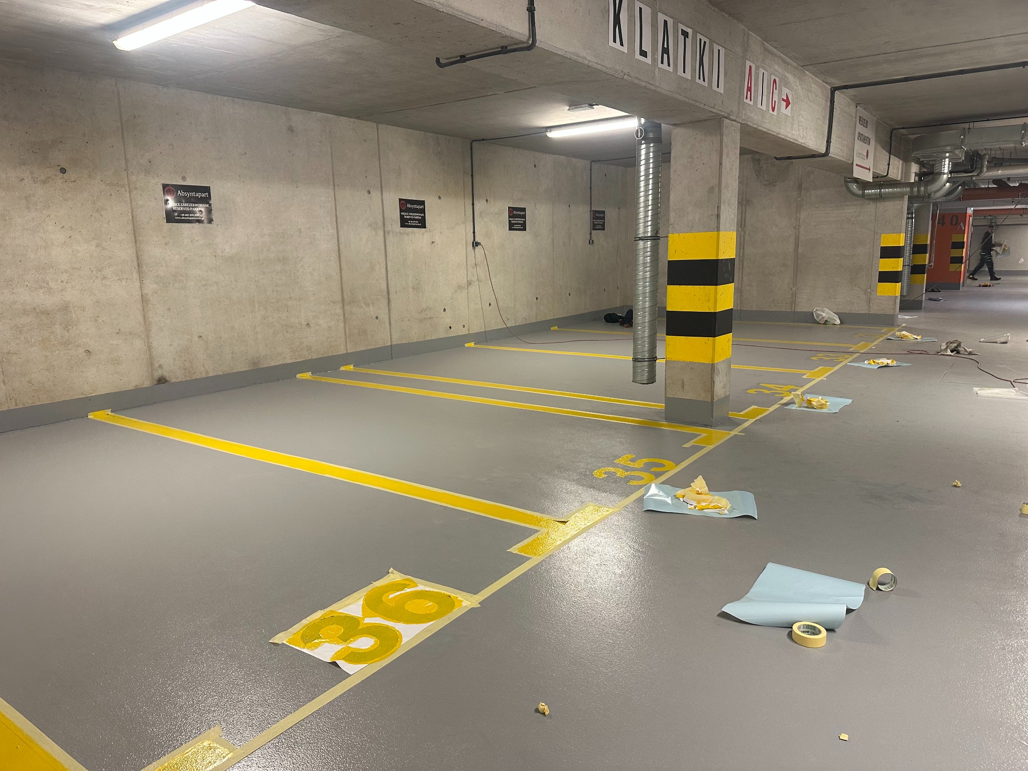 Oznakowane, szare miejsca parkingowe w garażu podziemnym, z żółtymi liniami i numerami, w trakcie malowania. Widoczne taśmy malarskie i szablony.