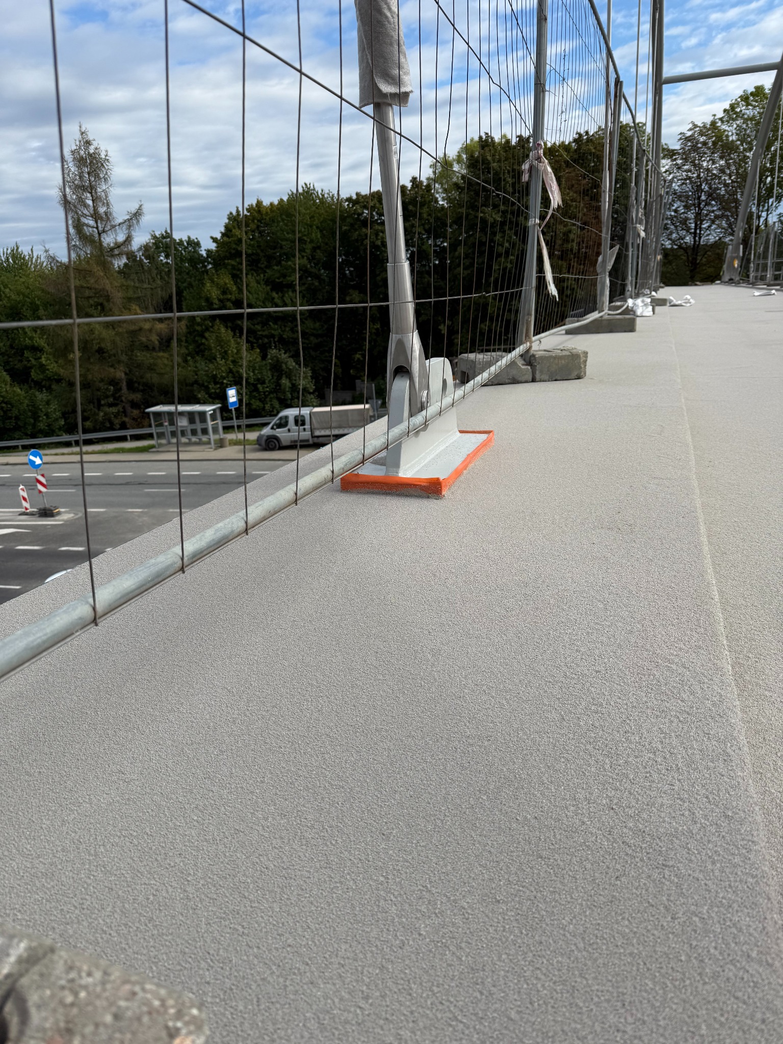 Szara, teksturowana powierzchnia z metalową barierką i widokiem na ulicę. Detal wykończenia posadzki na zewnątrz budynku z zabezpieczeniami.