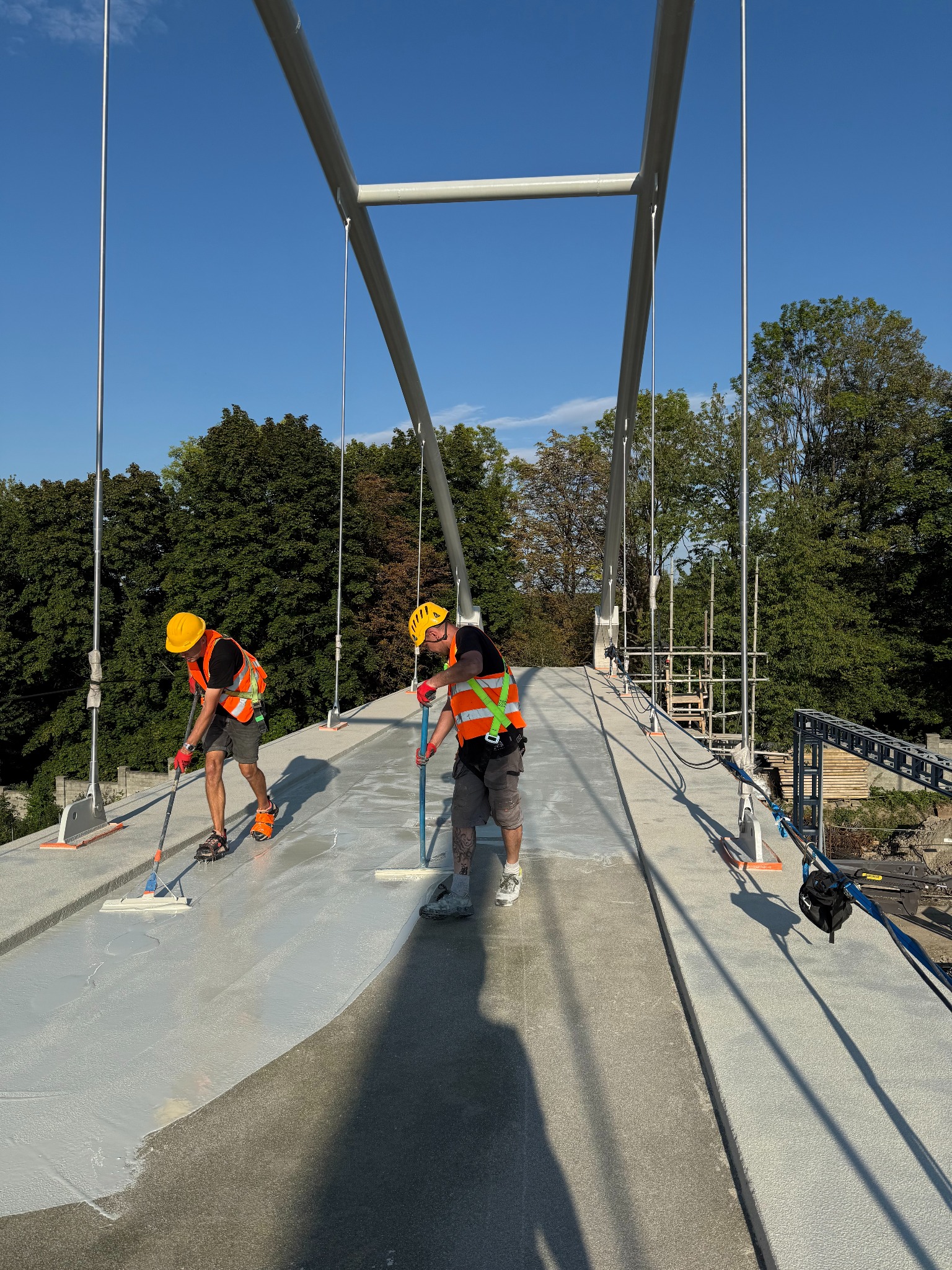 Dwóch robotników w kaskach i kamizelkach rozprowadza jasną substancję na betonowej powierzchni mostu, używając mopów. W tle zieleń drzew i konstrukcja mostu.